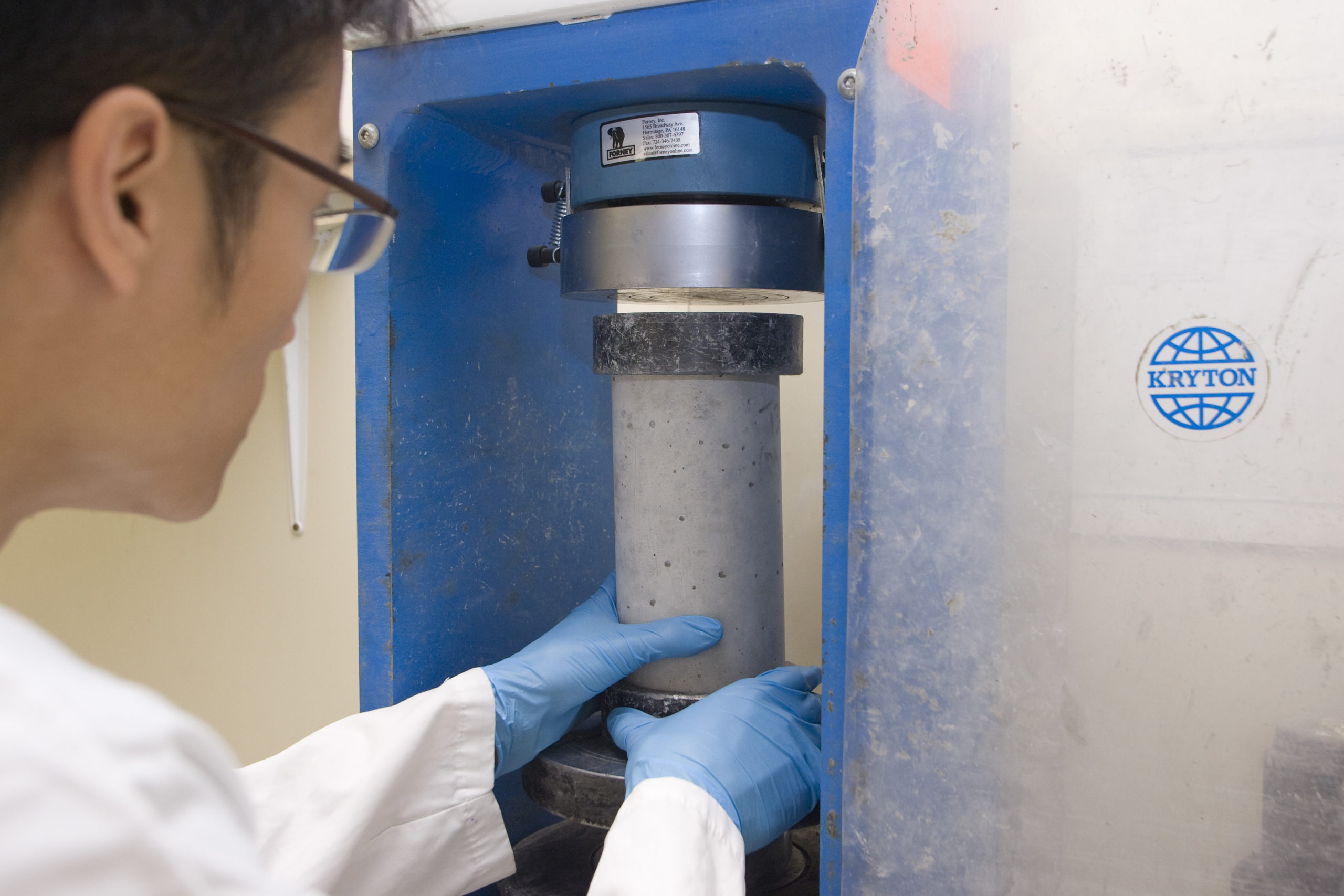A lab worker is placing a cylinder concrete sample in the compression strength test machine.